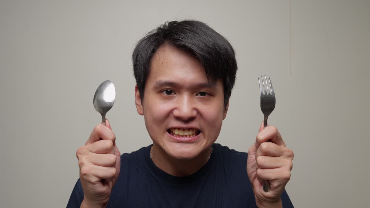 장난스럽고 탐욕스러운 표정이 젊은 아시아 남자의 얼굴을 가로질러 퍼져 나가고, 그는 꺼기와 포크를 움켜고, 그의 식사를 삼키기 위해 준비하고 있습니다.