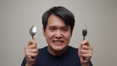 playful and ravenous look spreads across a young asian man's face as he clutches a spoon and fork, poised to devour his meal