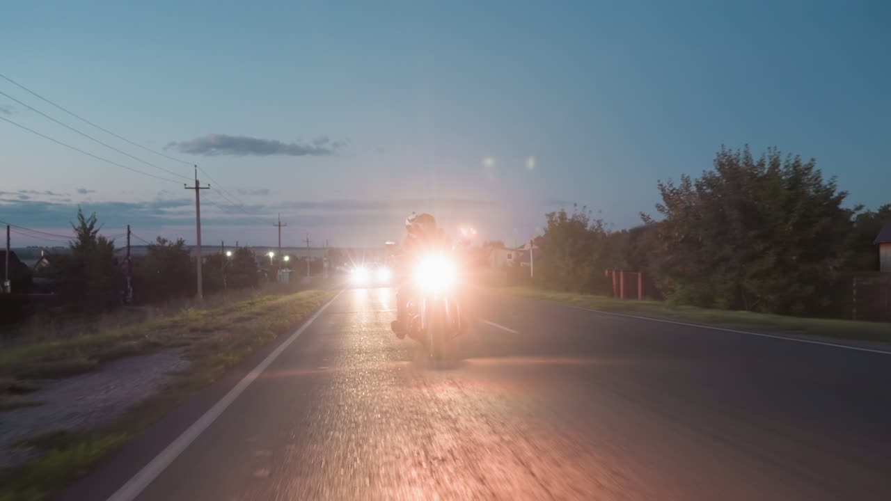 Biker riding skillfully at night along open road with glowing headlight reflecting across asphalt surrounded by houses, power poles and trees under fading blue evening sky with visible horizon