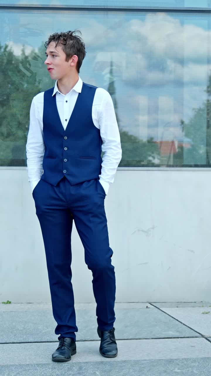 Giovane uomo con gilet blu, camicia bianca e pantaloni blu in piedi all'aperto, che sembra pensieroso mentre riflette sulle scelte di vita, ripresa verticale al rallentatore con telecamera fissa