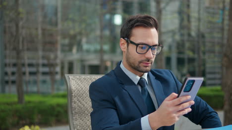 businessman picks up phone from the table and browsing internet, check emails, read news, shows smile in a park outdoors sitting in a chair