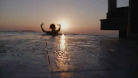 a woman silhouette dancing in infinity pool during sunset with sun reflections on wooden floor - defocused, slow motion, santorini, greece