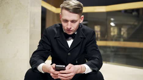 Nahaufnahme-Eines-Jungen-Blonden-Mannes-Im-Schwarzen-Anzug,-Der-Sein-Smartphone-Benutzt-Und-Auf-Der-Treppe-Im-Hotel-Sitzt