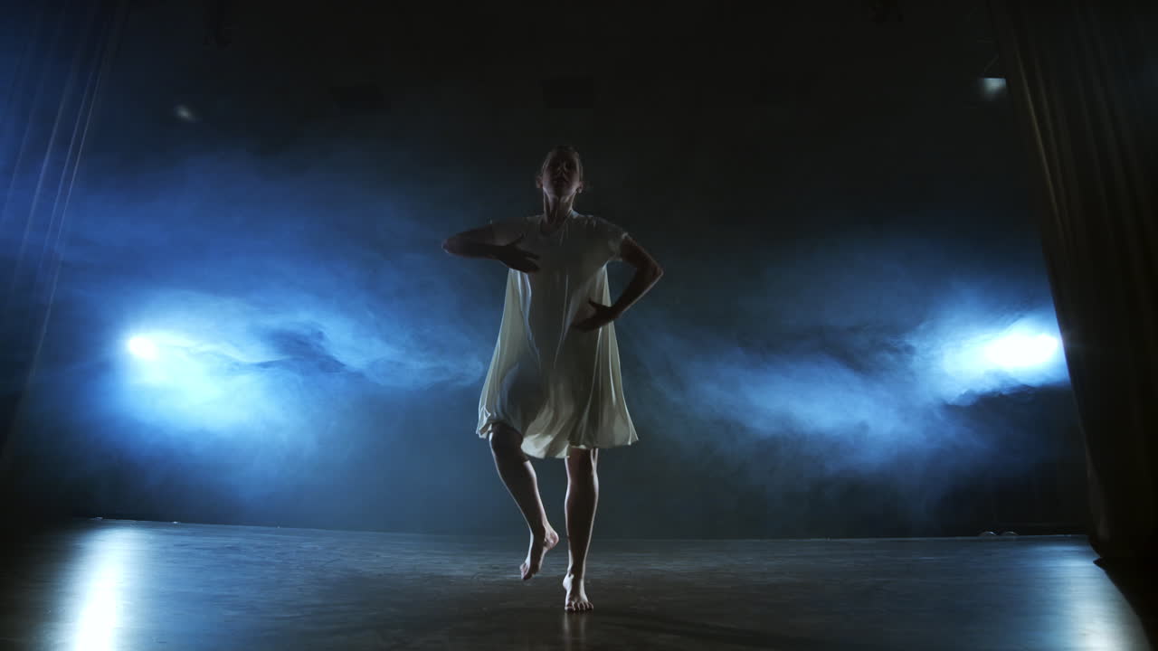 Modern dance woman in a white dress dances a modern ballet jumps on the stage with smoke in the blue spotlights.