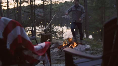 a man kneels by a campfire in the evening forest, adding logs to the fire