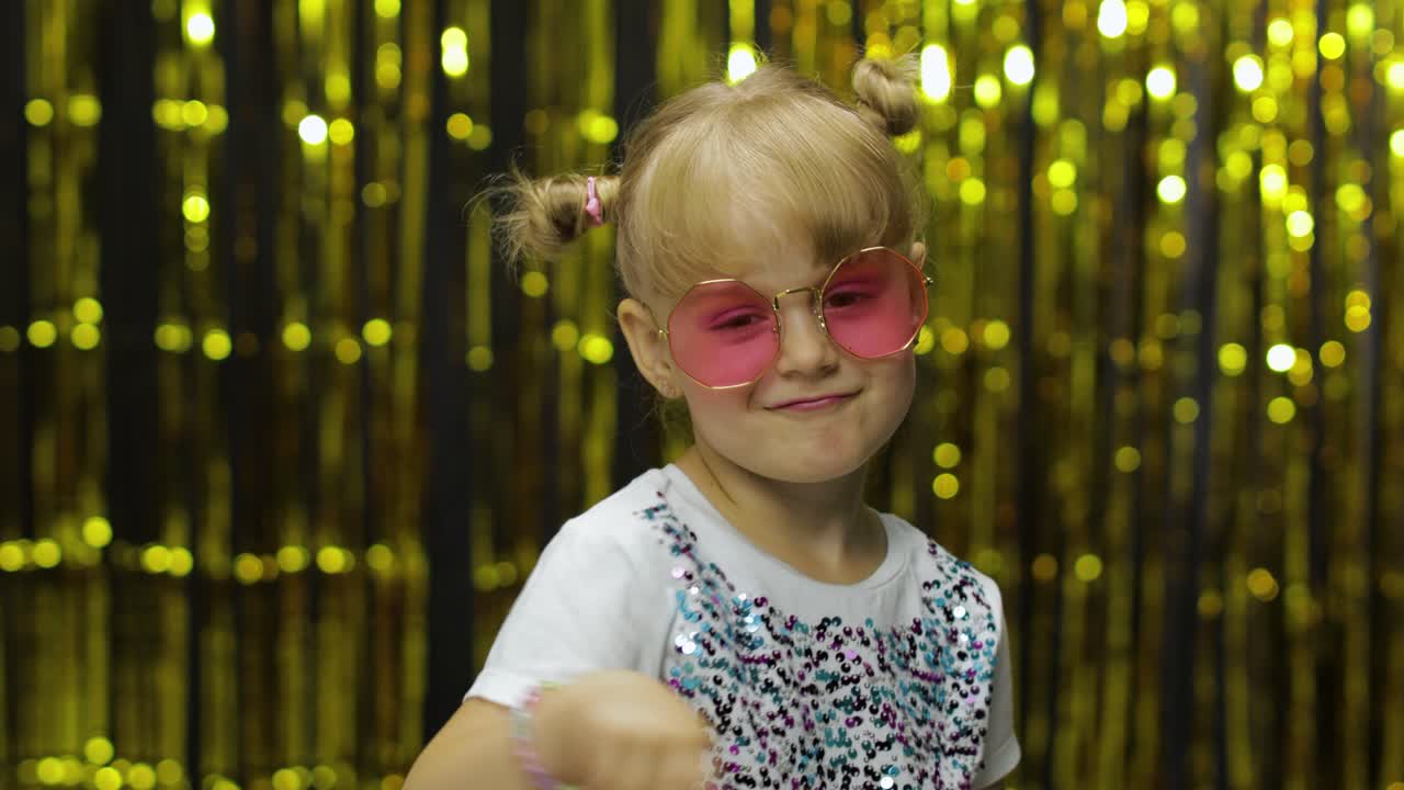 Child smiling, pointing fingers at camera. Girl posing on background with foil golden curtain