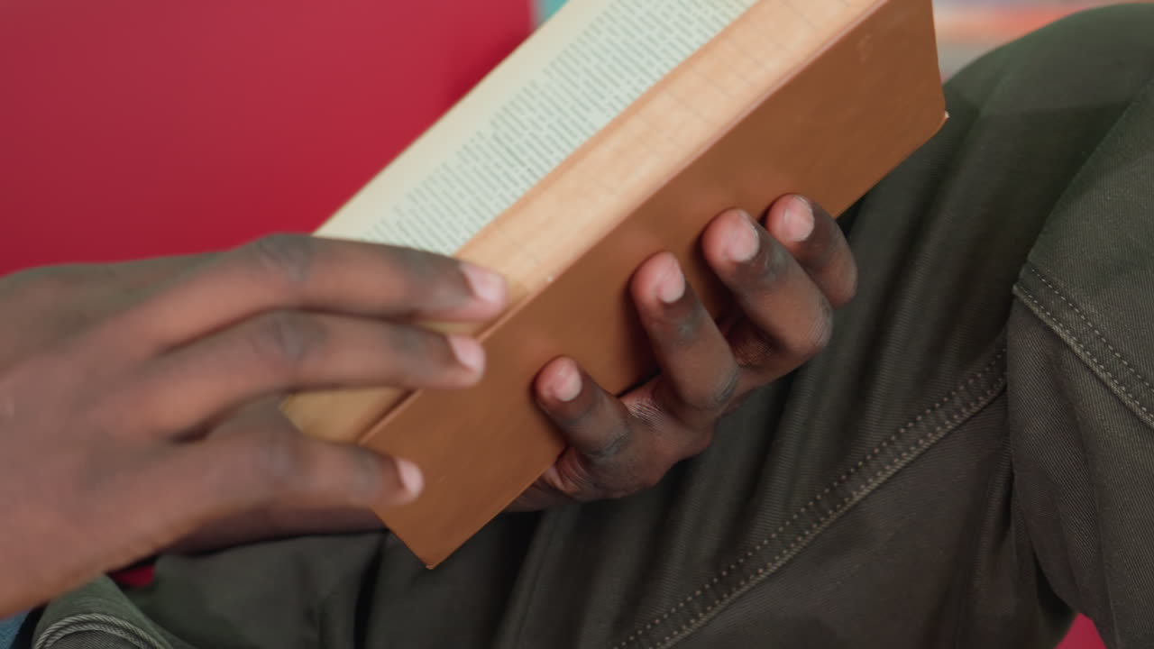 Primo piano di adulto che indossa una maglietta grigia e tiene in mano un libro con copertina rigida mentre è seduto su un divano rosso contro un muro color petrolio, concentrato sullo studio con la mano parzialmente visibile