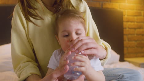 woman sitting with her baby on the bed in the bedroom while the baby playing with a feeding bottle