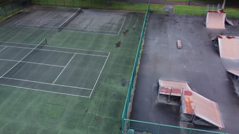 Aerial-view-flying-above-fenced-public-tennis-court-and-skate-park-ramp-in-empty-closed-playground