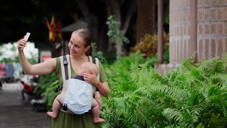 with her baby in a kangaroo backpack, a young woman captures memories on a mobile phone during her travels. she strolls and periodically looks at the phone screen