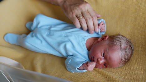 tiny newborn with blond hair wearing blue romper. baby lies in the crib while dad's hand caresses him. close up.
