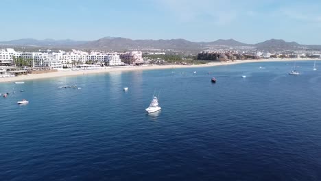 Aerial-view-of-the-beautiful-medano-beach-in-cabo-san-lucas-overlooking-the-blue-sea-with-boats-in-the-water,-hotel-facilities-and-majestic-mountains-in-the-background-on-a-sunny-day-on-vacation