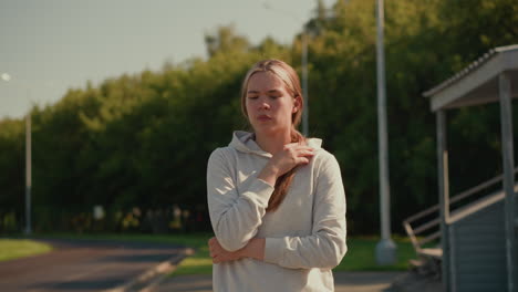 young woman in casual hoodie walking thoughtfully in empty stadium, gently touching her hair as warm sunlight illuminates trees in background