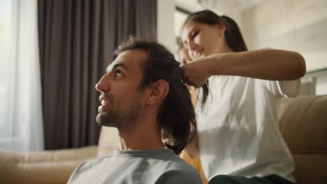 A-brunette-wife-in-a-white-T-shirt-braids-her-brunette-husband's-hair-while-sitting-on-a-light-brown-sofa-with-her-daughter-in-a-modern-room