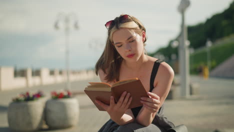 dama de solitario negro y gafas de sol descansando en su cabeza se sienta al aire libre, leyendo un libro cubierto de marrón pensativo como el viento levanta las páginas ligeramente, con un fondo borroso