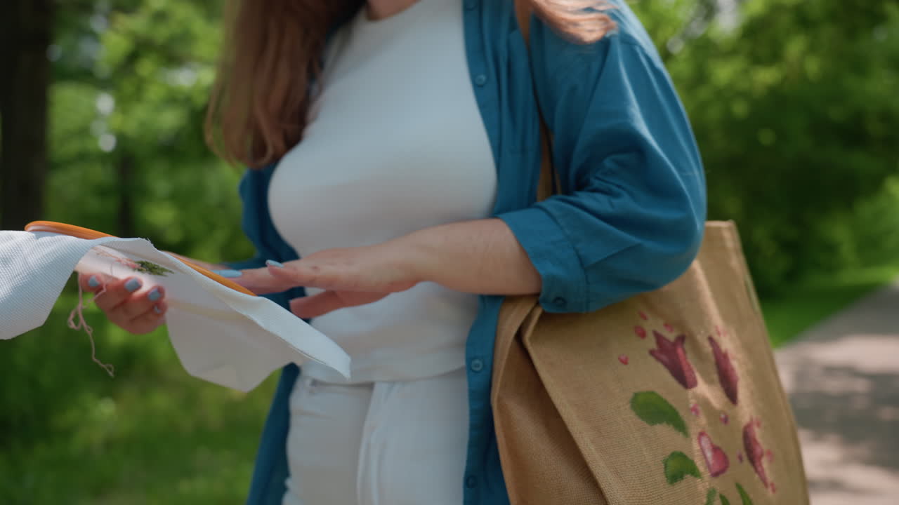Close torso view of woman in blue shirt dancing happily with embroidered fabric and hoop she created, showing blue manicure and canvas tote, sunshine and green trees in background