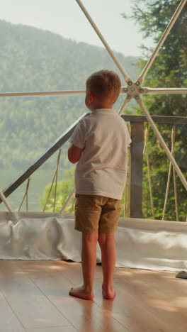 child stands alone under glass dome at glamping. thoughtful little boy looks out of panoramic window on terrace. kid observes calm nature scene