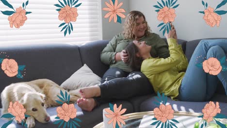 Animation-of-pink-flowers-over-happy-caucasian-lesbian-couple-relaxing-on-couch-using-smartphone