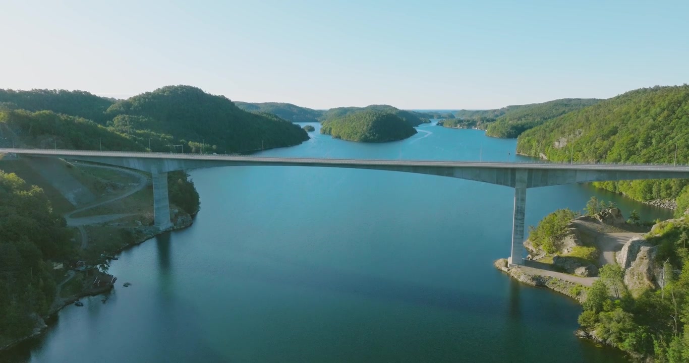 Luftfoto af bro, der krydser blåt fjordvand omgivet af skovklædte bakker i slowmotion