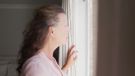 thoughtful senior caucasian woman standing in sunny room looking out of window