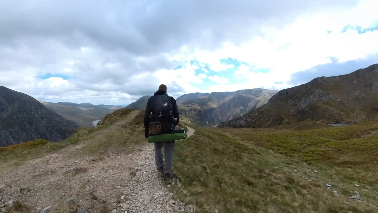 Hiker walks along rocky trail near Y Garn summit in Snowdonia with sweeping valley views below under cloudy sky, capturing solitude and adventure in scenic natural highlands of North Wales