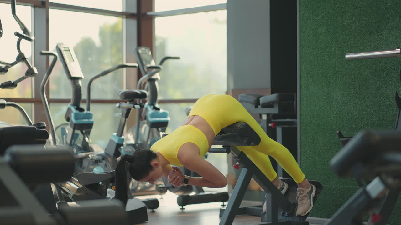 노란색 운동복을 입은 젊은 히스패닉 여성이 허리 근육을 훈련시키기 위해 과장 운동을 수행합니다. 갈색 여성은 허리와 자세를 강화하기 위해 체육관의 벤치에 몸을 들어 올립니다.