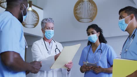 Diverse-group-of-male-and-female-doctors-in-face-masks-looking-at-files-and-discussing-in-hospital