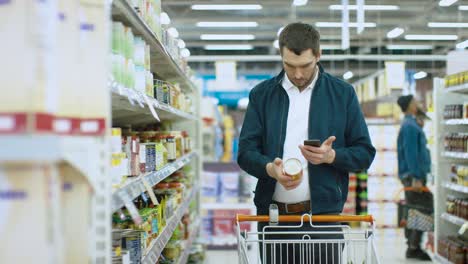 en el supermercado: un hombre guapo usa un teléfono inteligente y mira el valor nutricional de los productos enlatados. está de pie con el carrito de compras en la sección de productos enlatado.