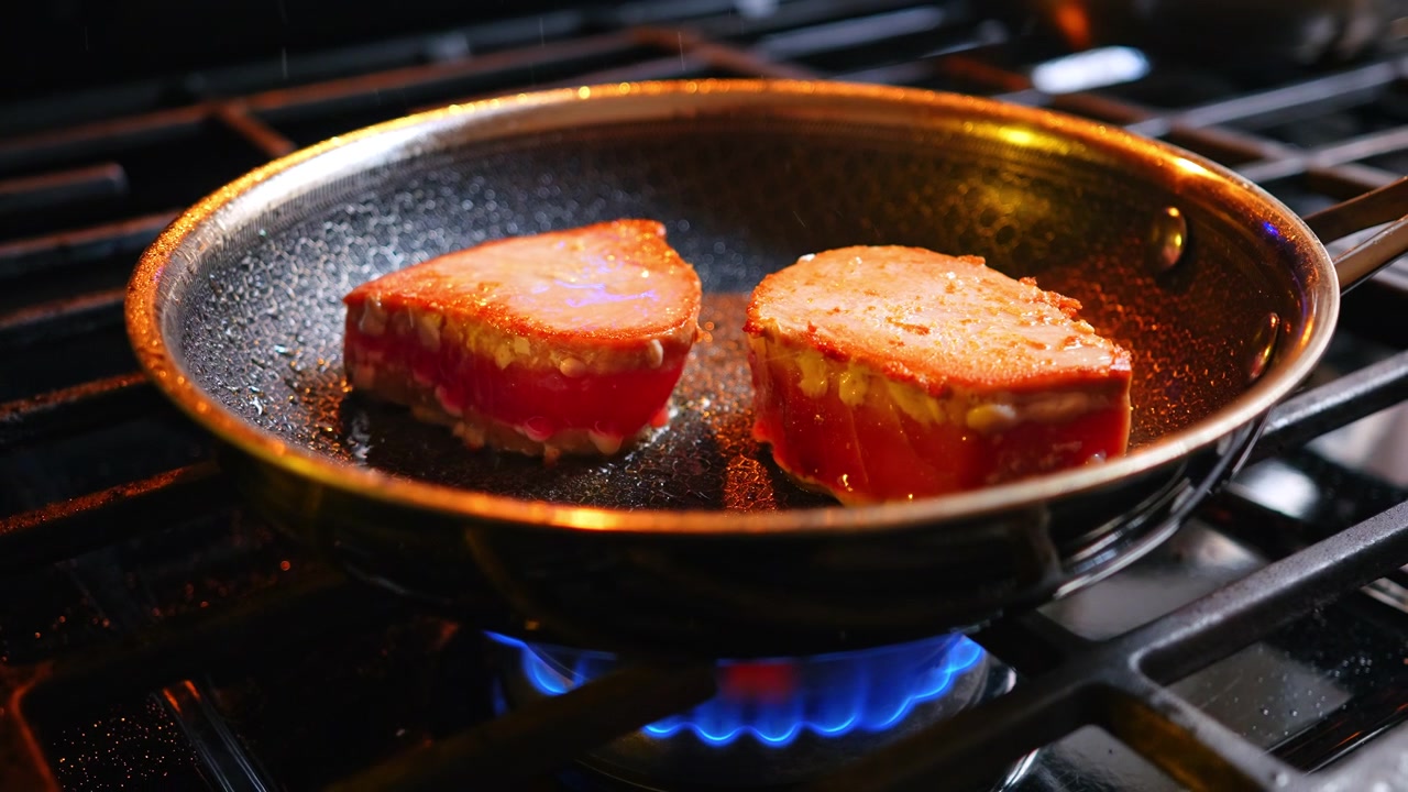 Tonfiskstekar som steks i en metallpanna över en blå gaslåga. Sidovy av två tonfiskfiléer som steks i en metallpanna med synliga blå gaslågor och ånga på en mörk spis.