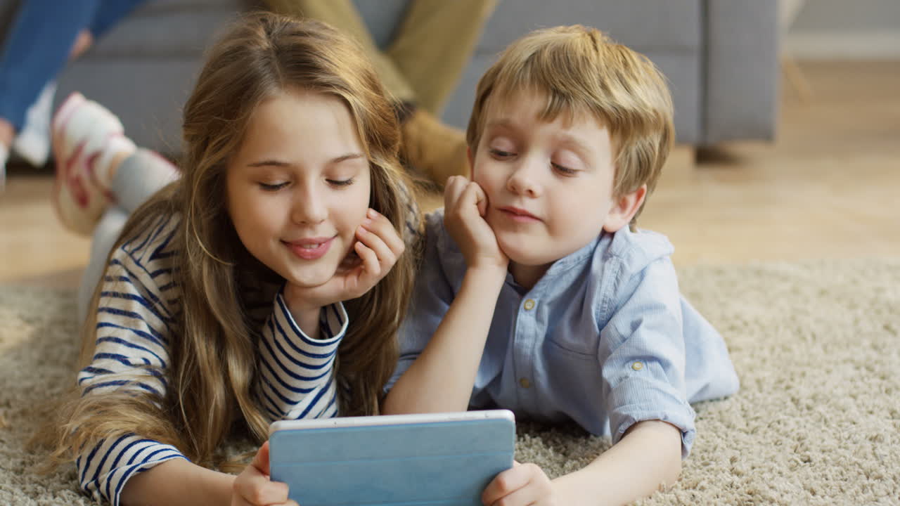 Close Up Of Two Cute Kids, Sister And Brother, Lying On The Carpet At Home And Watching Something Funny On The Tablet Computer