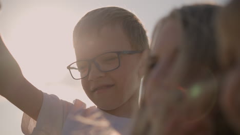 close-up of a young boy wearing glasses and a white shirt, standing beside his blurred mother. the boy points at the sky, his expression focused, capturing a curious and thoughtful moment outdoors