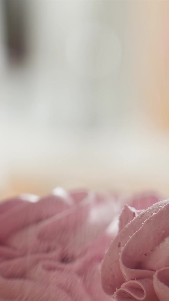 Primo piano di decorazione di glassa rosa con texture liscia. Particelle bianche cadono sulla superficie glassata, coprendola gradualmente sempre di più. L'illuminazione soffusa crea uno sfondo sfocato, enfatizzando.
