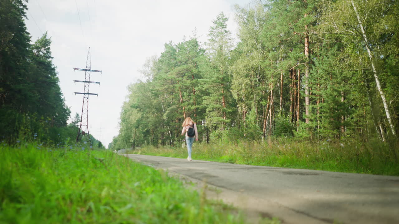 Close up of wild greenery and puddle on rural roadside with woman walking in distance under overcast sky, surrounded by forest and electric poles stretching into horizon
