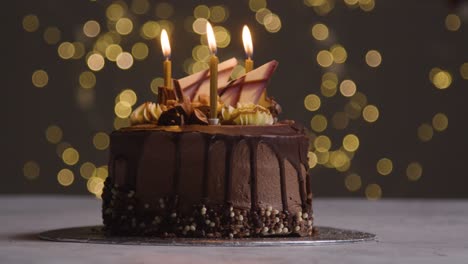 Foto-De-Estudio-De-Pastel-De-Celebración-De-Cumpleaños-De-Chocolate-Decorado-Con-Velas-Encendidas-Contra-La-Iluminación-De-Fondo-Del-Bokeh