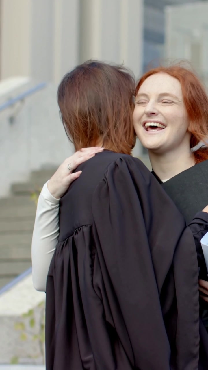 Video verticale: Fine della cerimonia di laurea, due donne in toga nera si abbracciano, prendendo il diploma da una cartella sui gradini