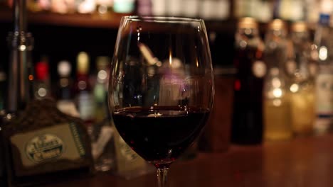 close-up of a red wine glass on a wooden bar with blurred bottles in the background.