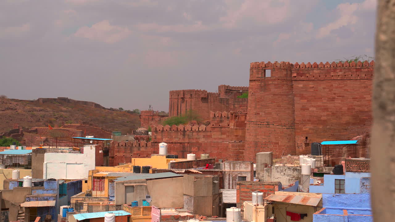 Una ripresa statica delle imponenti mura difensive in arenaria rossa del Forte Mehrangarh che si ergono sopra gli edifici residenziali colorati e i tetti della Città Blu a Jodhpur, Rajasthan, India.
