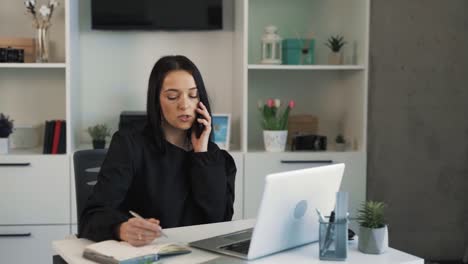 una mujer de negocios ocupada se sienta en un escritorio en la oficina y habla por teléfono, resuelve asuntos de trabajo y escribe en un cuaderno