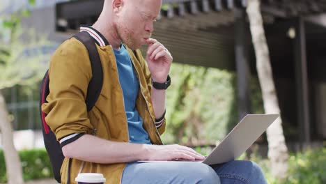 hombre albino afroamericano pensativo con rastas sentado en el parque usando una computadora portátil