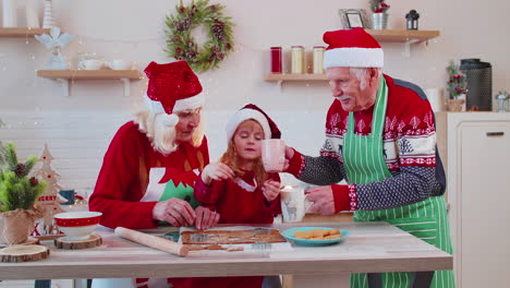 Senior-man-with-cups-hot-chocolate-walking-into-Christmas-home-kitchen-to-grandmother-and-grandchild