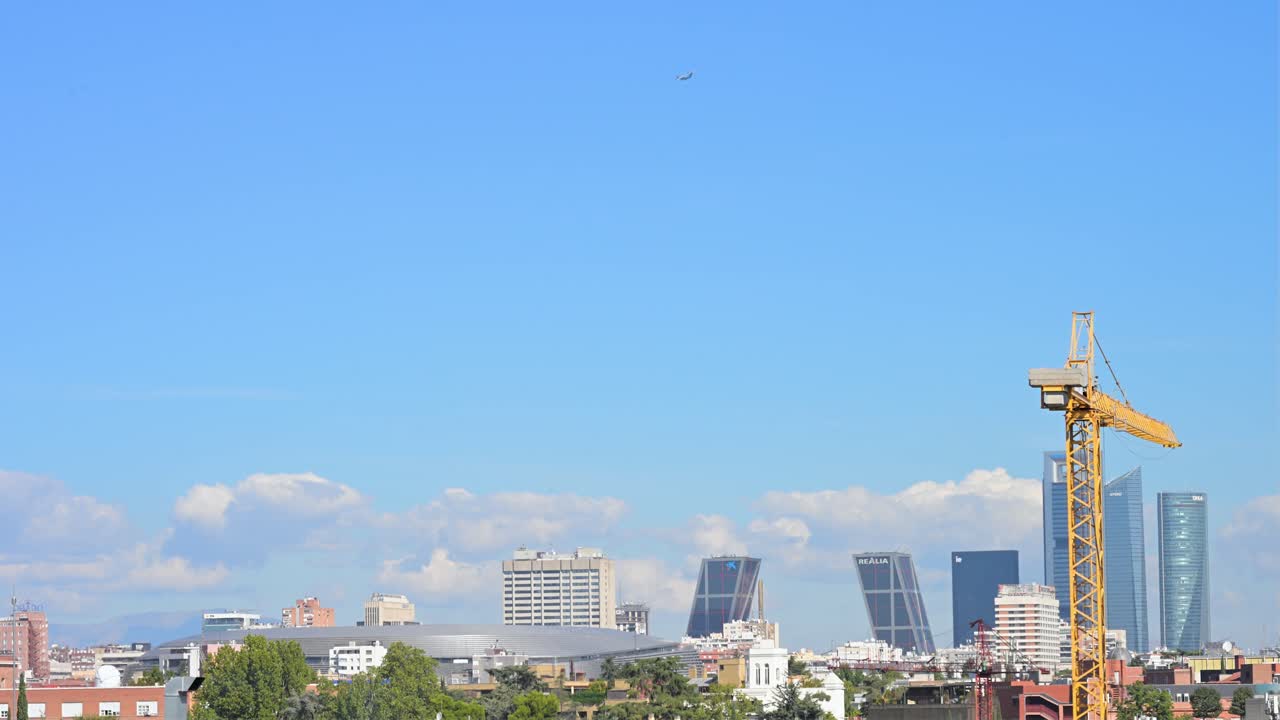 A construction crane frames the Madrid skyline, a symbol of urban development. The view features the iconic KIO Towers and Cuatro Torres skyscrapers as a plane flies across the clear blue sky