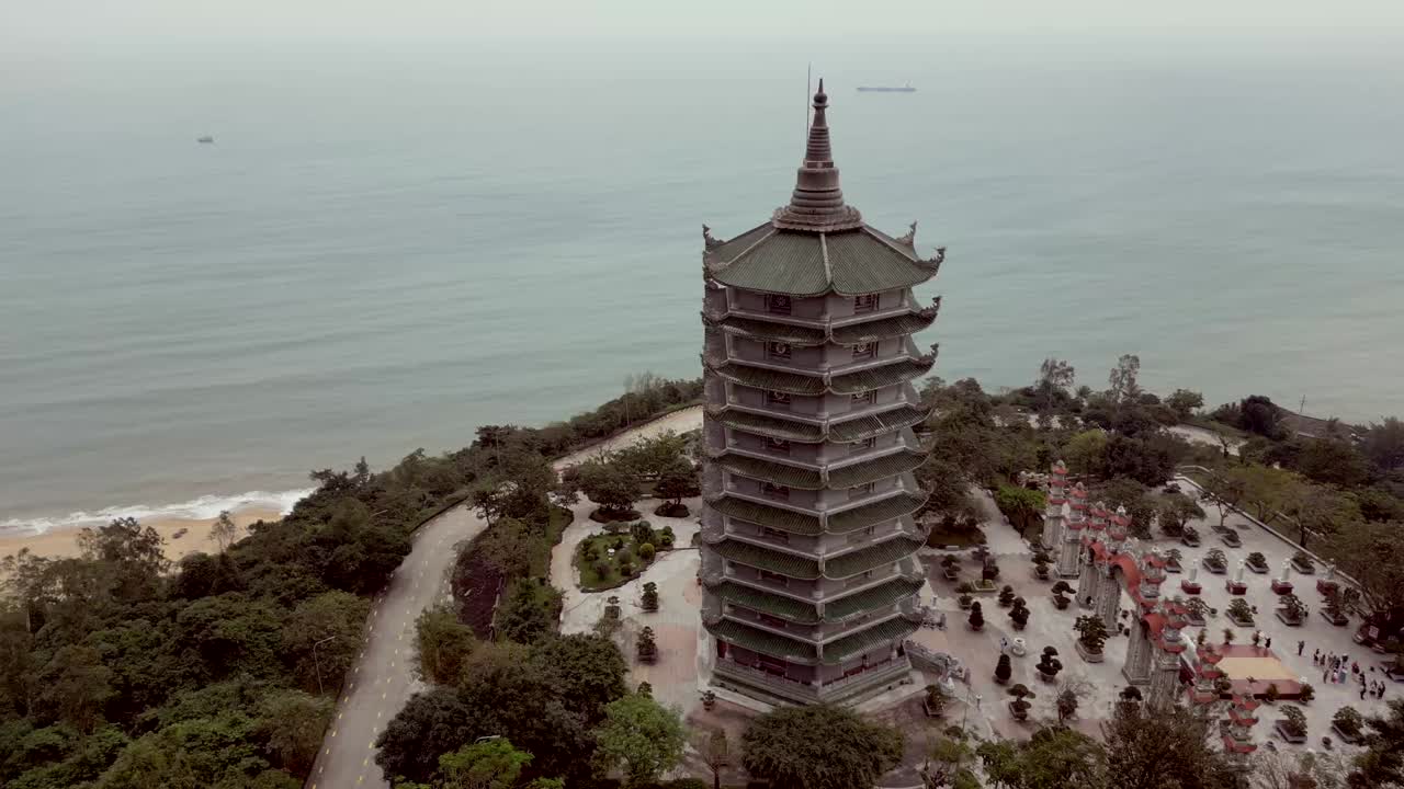 Drone view of a tall, multi-level pagoda near a calm sea, surrounded by trees, paths, gardens, and distant coastline under soft daylight.