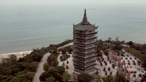 drone view of a tall, multi-level pagoda near a calm sea, surrounded by trees, paths, gardens, and distant coastline under soft daylight.