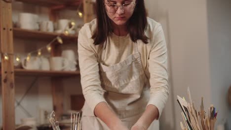 young woman potter in glasses kneads clay on the table in art studio