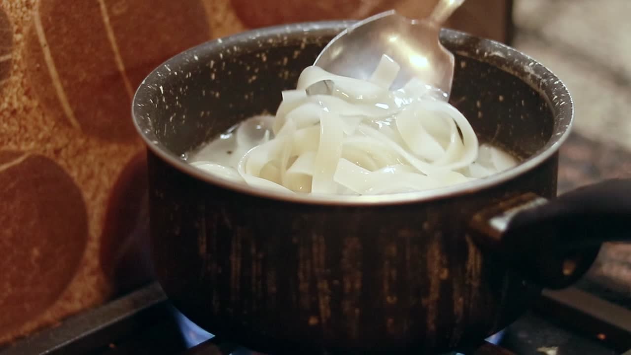 Stirring flat rice noodles known as Kuey Teow, Kuay Tiew, or Hor Fun boiling in a pot on a stove in preparation for cooking stir-fry dish