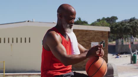 Hombre-Afroamericano-Mayor-Con-Baloncesto-Usando-Un-Teléfono-Inteligente-Cerca-De-La-Playa