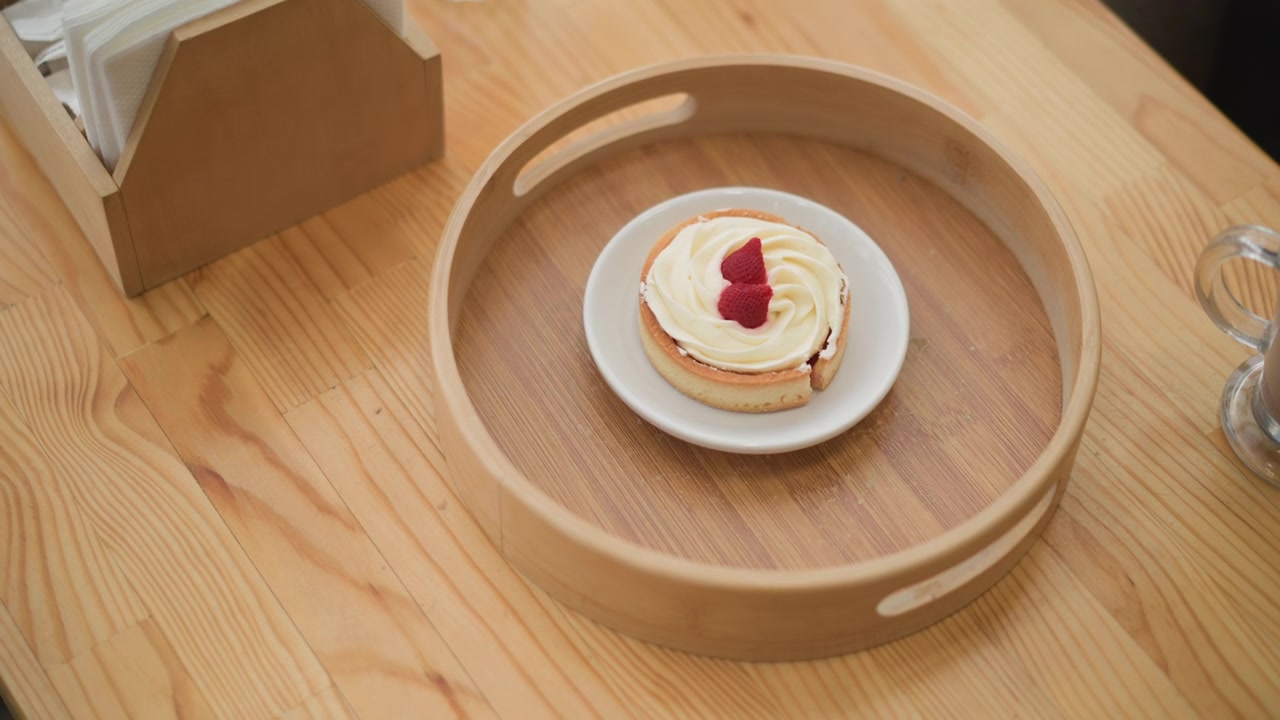 Layered dessert topped with strawberry cream garnish rests on white plate inside round wooden serving tray, set on light wood table with soft natural light and clean cozy presentation
