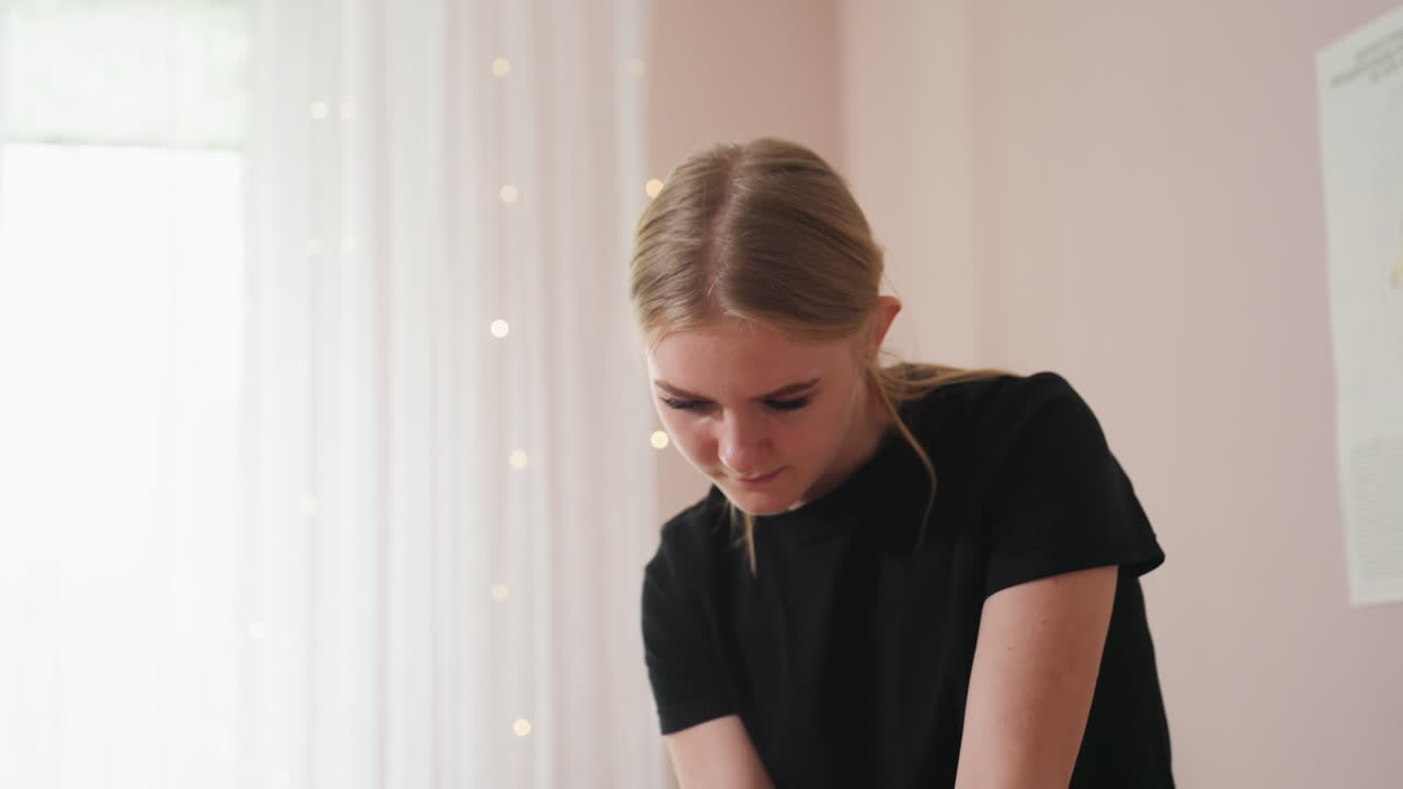Light skin blonde woman wearing black shirt looking downward in softly lit room, white sheer curtain in background and blurred anatomy poster on wall add professional context to calm setting