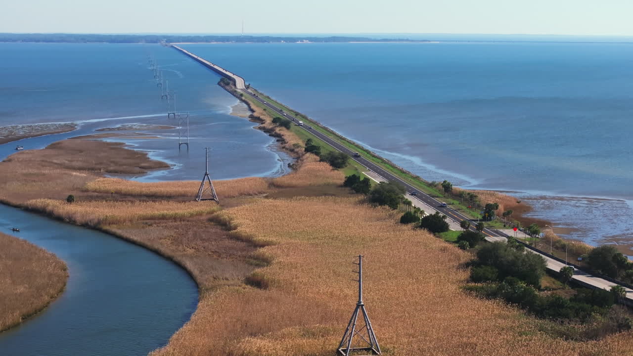Eine Luftaufnahme im Schwenk erfasst die John Gorrie Memorial Bridge, die sich über das schimmernde türkisfarbene Wasser der Apalachicola Bay in Florida erstreckt. Als Hauptverbindung auf dem US Highway 98 verbindet die Brücke…