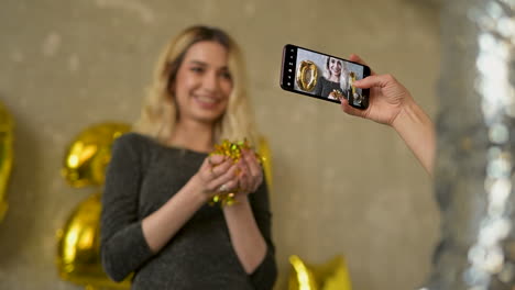 hands taking a photo of a young pretty woman blowing gold glitter confetti and wearing evening dress on new year's eve